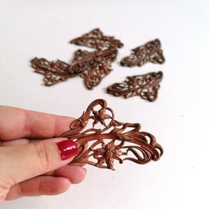 May include: A set of eight ornate, copper-colored metal embellishments with a floral design. The embellishments are intricate and detailed, with a delicate, filigree-like appearance.