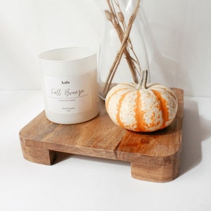 May include: A white candle with the text "kala Fall Breeze Scented Soy Candle Hand Poured 9 oz" on the label, a small orange and white pumpkin, and dried branches in a glass vase on a wooden tray.