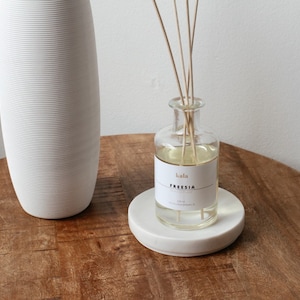 May include: A white, ribbed ceramic vase and a glass aromatherapy diffuser with reeds on a wooden table. The diffuser has a label that reads "kala FREESIA". The diffuser sits on a white marble coaster. The table has a natural wood grain.