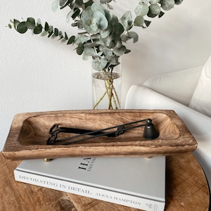May include: A wooden, rectangular tray with a carved interior holds black candle accessories. The tray sits atop a book titled "Decorating in Detail" on a round, wooden table. A glass vase with eucalyptus branches is in the background.