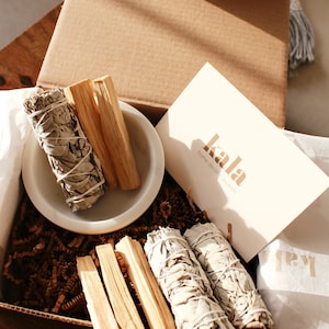 May include: A brown cardboard box filled with white sage bundles and palo santo sticks. The box is decorated with brown paper shreds and a white card with the text "kala home decor accents".