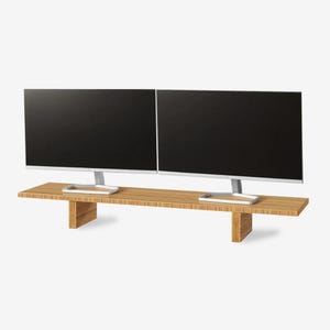 May include: Two black computer monitors with silver stands sit on a light brown wooden monitor stand. The stand has a rectangular top and two supporting legs. The monitors are side-by-side.