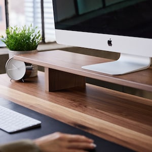 May include: A wooden desk with a monitor stand. The stand is made of light brown wood and supports a white computer monitor. A small potted plant and a silver alarm clock are on the desk. The desk has a two-tone wood grain.