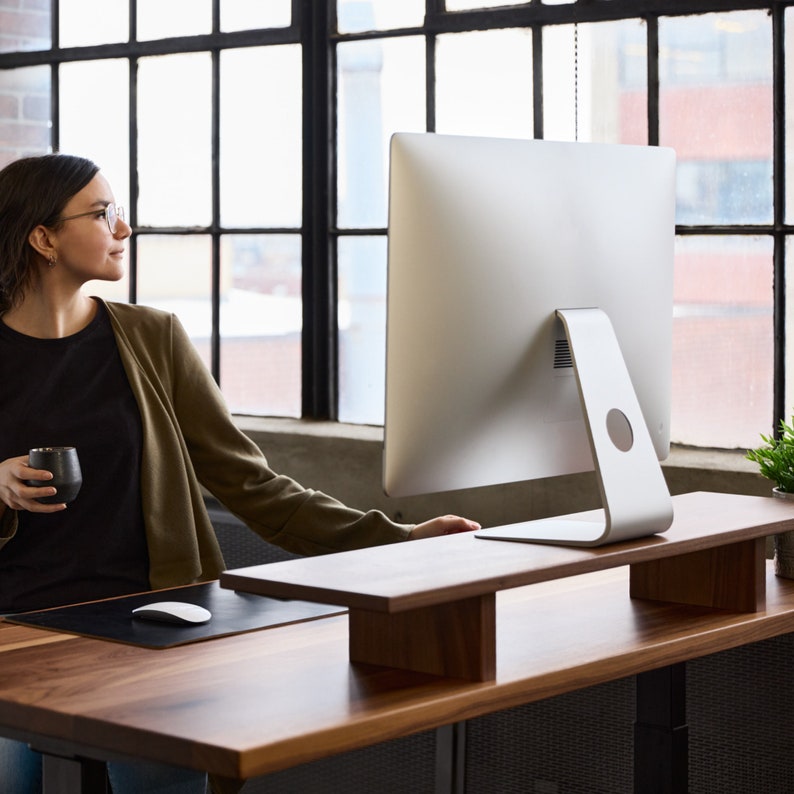May include: A wooden desk with a computer monitor and a wooden monitor stand. A black mouse and mousepad are on the desk. A person is holding a black mug. The background includes a large window.