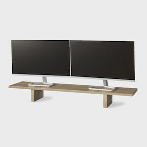 May include: Two black computer monitors on a light brown wooden desk. The desk has a rectangular top and two supporting legs. The monitors have silver stands.