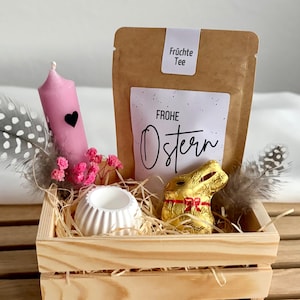 Könnte beinhalten: Eine Holzkiste mit Osterartikeln. Sie enthält eine rosa Kerze mit schwarzem Herz, einen Teebeutel, einen goldverpackten Schokoladenhasen, einen weißen Keramikhalter sowie dekorative Federn und Blumen. Auf dem Teebeutel steht "Frohe Ostern".