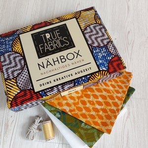 Könnte beinhalten: Eine farbenfrohe Box mit dem Text "TRUE FABRICS NÄHBOX NACHHALTIGES NÄHEN DEINE KREATIVE AUSZEIT" auf der Vorderseite. Die Box ist geöffnet und enthält Stoff, Garn und Schnur.