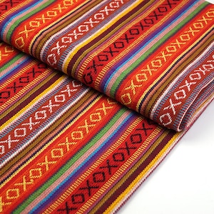 Op de afbeelding: Gevouwen stof met een levendig gestreept patroon. Het ontwerp heeft afwisselende banden van rood, oranje, geel, groen, blauw en paars, met een herhaald wit geometrisch motief. De stof lijkt geweven.