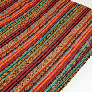 Op de afbeelding: Een kleurrijk geweven textiel met een gestreept patroon. Het ontwerp heeft afwisselende banden van rood, oranje, turquoise en zwart, met decoratieve ruitpatronen. De stof is gevouwen en toont de textuur en details van het weefsel.