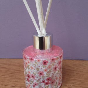 May include: A floral reed diffuser. The glass bottle is decorated with a pink floral pattern and a silver cap. The diffuser contains white reeds. The bottle is approximately 10 cm tall.
