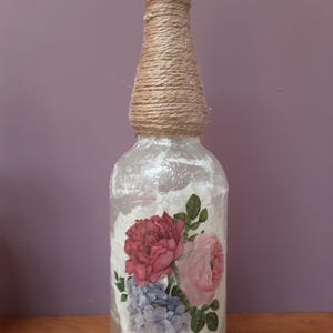 May include: A clear glass bottle decorated with a floral design of pink and blue flowers. The neck is wrapped in natural twine, and it has a brown stopper. The bottle is a decorative item.
