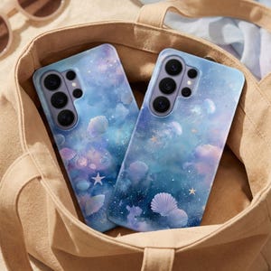May include: Two phone cases with a dreamy, ocean-themed design featuring seashells, stars, and a galaxy-like background in shades of blue, purple, and white. The cases are inside a beige tote bag, with sunglasses and seashells scattered around.
