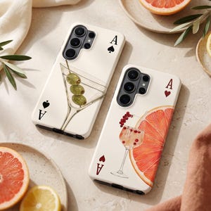 May include: Two cream-coloured phone cases with playing card designs. One shows a martini glass with olives, the other a cocktail with a grapefruit slice. Both cases have an Ace of Spades or Hearts.