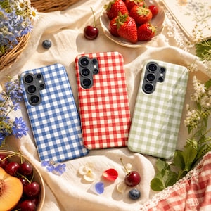 Peut inclure: Trois coques de smartphone avec un motif à carreaux vichy bleu, rouge et vert clair. Chaque coque présente une découpe pour appareil photo multi-objectifs dans le coin supérieur gauche, disposées sur un tissu blanc avec des fruits éparpillés.