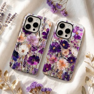 Peut inclure: Deux coques de téléphone avec un motif floral composé de fleurs violettes, blanches et pêche. Les coques ont une finition brillante et une découpe noire pour l'appareil photo. Le fond est un tissu blanc avec des brins de lavande et des feuilles dorées.