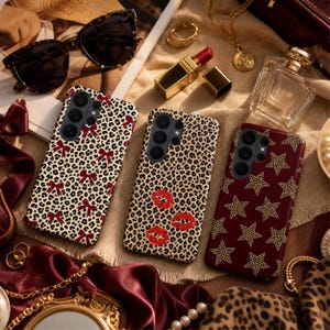 Peut inclure: Trois coques de smartphone aux motifs distincts : une avec un imprimé léopard et des nœuds rouges, une avec un imprimé léopard et des lèvres rouges, et une avec un fond rouge foncé ornée d'étoiles à motif léopard aux contours dorés.