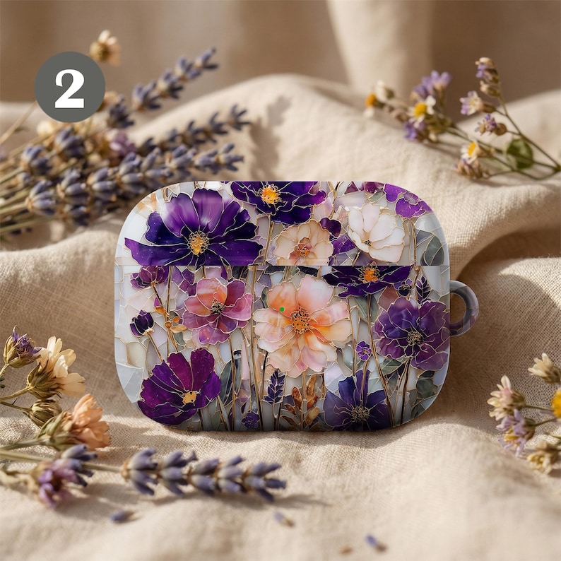 May include: A case for wireless earbuds, decorated with a floral pattern. The design features purple, pink, and white flowers in a mosaic style. The background is a neutral beige, and the case has a small loop for attachment.