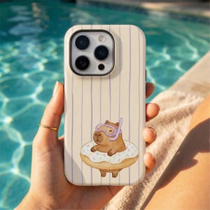 May include: A phone case with a cream background and vertical purple stripes. The case features a cartoon capybara wearing a snorkel and floating on a donut. The phone case is held by a person with a gold ring.