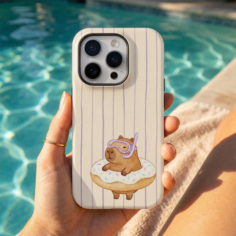May include: A phone case with a cream background and vertical purple stripes. The case features a cartoon capybara wearing a snorkel and floating on a donut. The phone case is held by a person with a gold ring.