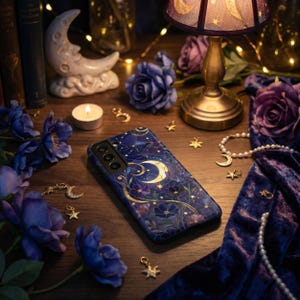May include: A dark blue phone case with a celestial design featuring a crescent moon, stars, and floral accents. The case is surrounded by purple roses, gold charms, and a small lamp with a moon and star pattern.
