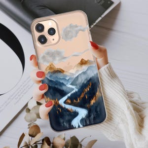 Puede incluir: Una funda transparente para teléfono con un diseño de acuarela de una cordillera y un río que serpentea a través de ella. Las montañas están pintadas en tonos de azul, marrón y dorado, con nubes blancas en el cielo.