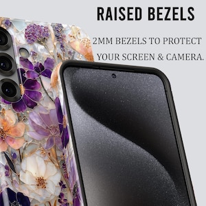 May include: A smartphone case with a floral design in shades of purple, white, and gold. The case has raised bezels, with the text "RAISED BEZELS" and "2MM BEZELS TO PROTECT YOUR SCREEN & CAMERA."