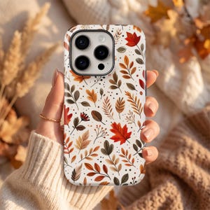 Puede incluir: Una funda para smartphone con fondo color crema que presenta un patr&oacute;n repetitivo de hojas de oto&ntilde;o en tonos rojo, naranja, marr&oacute;n y verde oscuro, junto con peque&ntilde;os puntos negros e ilustraciones de ramas delicadas en toda la superficie.