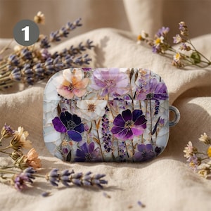 May include: A case for wireless earbuds with a floral design. The case features purple, white, and peach-coloured flowers with gold accents. The background is light grey, and the case has a small loop on the side.