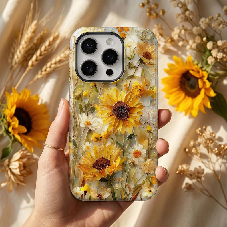May include: A floral phone case featuring a vibrant design of yellow sunflowers and white daisies. The case has a glossy finish and a detailed, artistic rendering of the flowers. The background is a soft, neutral color, enhancing the bright floral pattern.