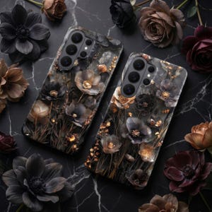 Op de afbeelding: Twee telefoonhoesjes met een bloemenontwerp met donkergrijze, zwarte en gouden bloemen. De hoesjes zijn omgeven door donkere rozen en andere bloemen op een zwart marmeren oppervlak. De hoesjes hebben meerdere cameralenzen.