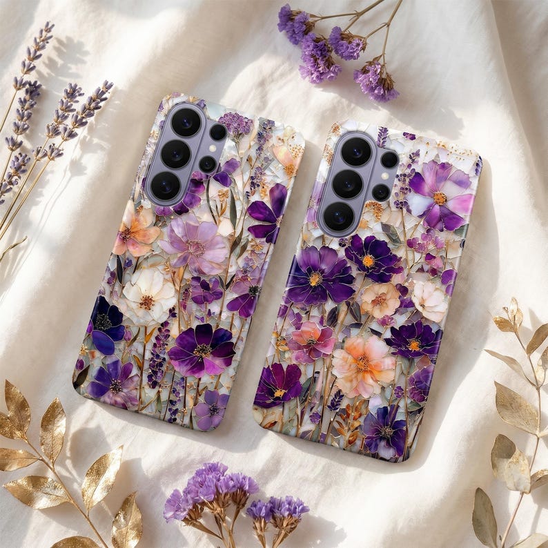 May include: Two phone cases with a floral design. The cases feature a variety of flowers in shades of purple, white, and orange, set against a white background. The cases have a glossy finish and are decorated with gold accents.