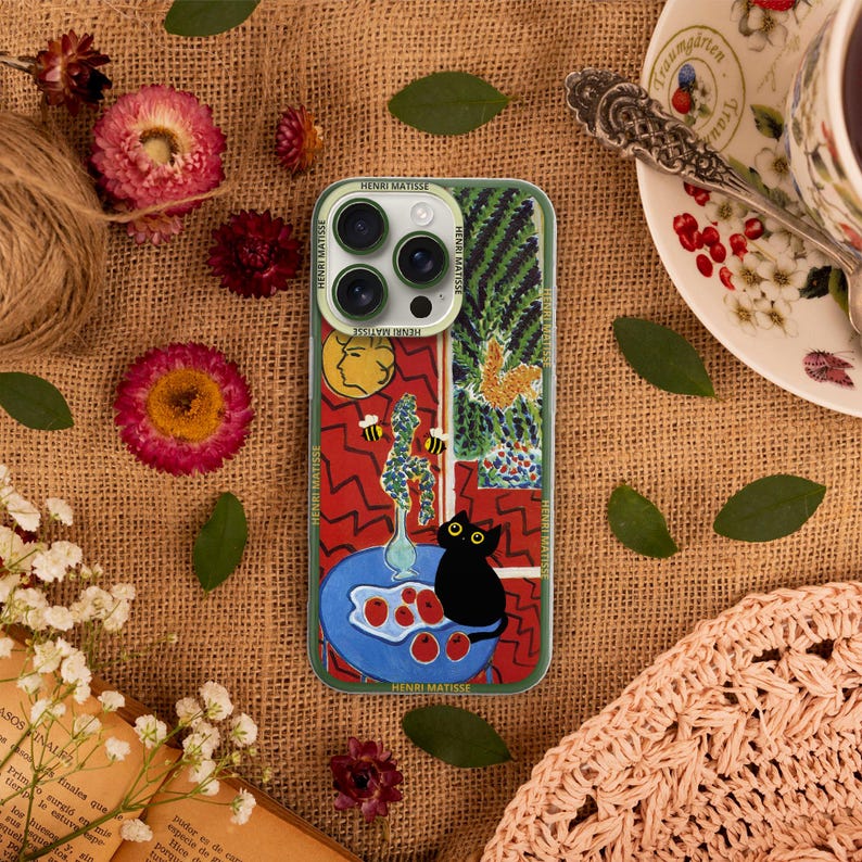 K&ouml;nnte beinhalten: Eine Handyh&uuml;lle mit einem farbenfrohen abstrakten Design, das eine schwarze Katze, rote &Auml;pfel und eine Vase mit Blumen zeigt. Die H&uuml;lle hat einen Ausschnitt f&uuml;r das Kameraobjektiv und der Text "HENRI MATISSE" ist unten aufgedruckt.