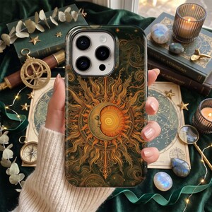 May include: A dark green smartphone case featuring a detailed gold and orange illustration of a crescent moon and a sun with swirling rays. The design includes intricate linework and small star accents across the entire surface of the case.
