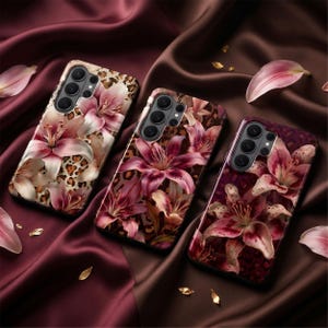 May include: Three phone cases with floral designs. Each case features a different color scheme of pink lilies and a leopard print background. The cases are displayed on a dark burgundy satin fabric, with scattered petals and gold accents.
