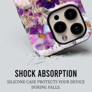 May include: A close-up of a phone case with a floral design in shades of purple, pink, and white. The case has a glossy finish and a black camera module. The text "SHOCK ABSORPTION" and "SILICONE CASE PROTECTS YOUR DEVICE DURING FALLS" are visible.