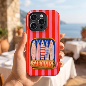 May include: A red and pink striped phone case featuring a sardine can design. The sardine can illustration includes four colorful fish and the word "SARDINES" in gold lettering. The phone case is held in a person's hand.