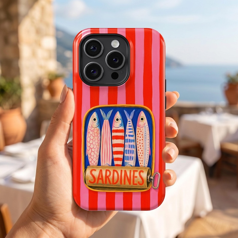 May include: A red and pink striped phone case featuring a sardine can design. The sardine can illustration includes four colorful fish and the word "SARDINES" in gold lettering. The phone case is held in a person's hand.