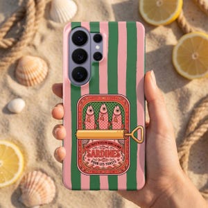 May include: A phone case with a pink and green striped design. The case displays a sardine can graphic with the text "Tout petit poisson dans la mer" and "Sardines pour les toasts". The sardine can graphic is red and gold in colour.