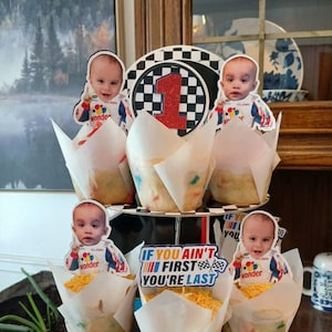 May include: A tiered cupcake stand with six cupcakes, each topped with a baby's photo and a racing-themed design. The cupcakes are decorated with shredded cheese. The stand has a checkered pattern and the text "If you ain't first you're last".