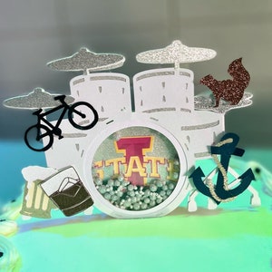 May include: A multi-layered cake topper with a drum set design. The topper includes a bicycle, squirrel, anchor, and beer mugs. The center features the word "STATE" in a circular frame. The cymbals and squirrel are glittery.