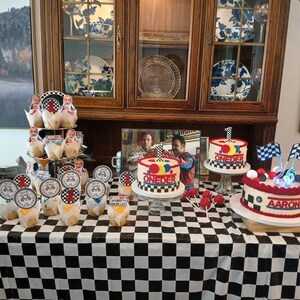 May include: A birthday party table with a black and white checkered tablecloth. The table is decorated with cupcakes, cake pops, and three cakes. The cakes are decorated with racing flags and the word "ONEDER" and "Aaron".