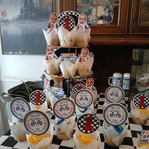 May include: Cupcakes with racing-themed decorations. Each cupcake has a paper wrapper and is topped with a circular label featuring a checkered flag and the number 1. Some labels read "Elias is the best there is".