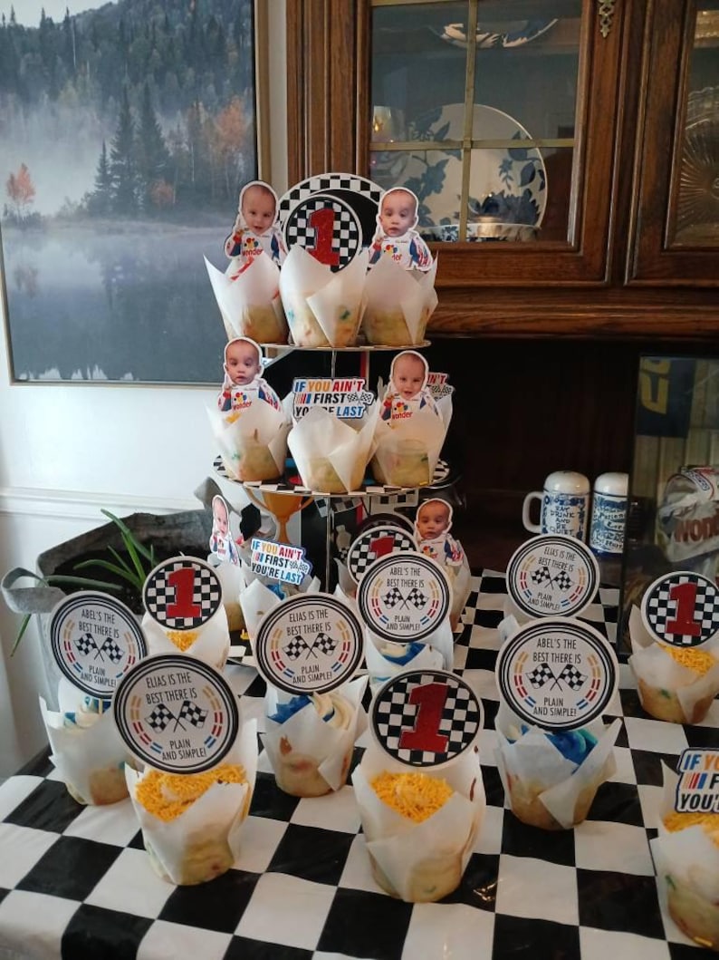 May include: Cupcakes with racing-themed decorations. Each cupcake has a paper wrapper and is topped with a circular label featuring a checkered flag and the number 1. Some labels read "Elias is the best there is".
