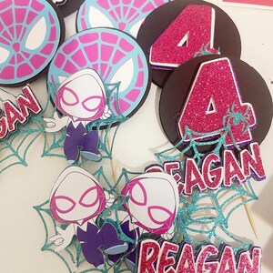 May include: A collection of superhero-themed party decorations. The set includes black circular toppers with a pink glitter number 4, and teal spiderwebs. Also included are cartoon character cutouts and name tags that read "REAGAN".