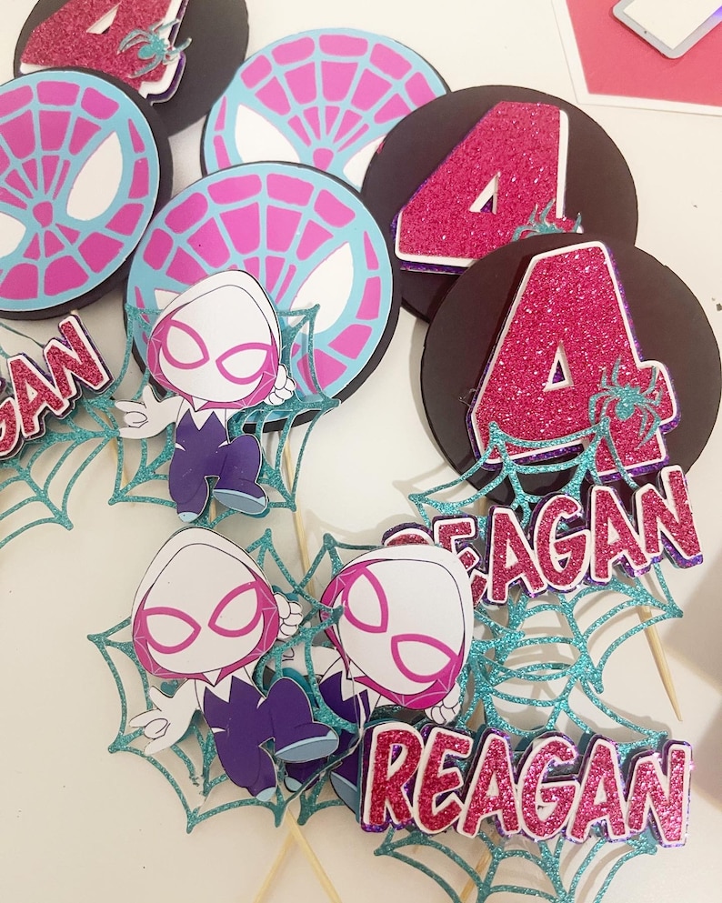 May include: A collection of superhero-themed party decorations. The set includes black circular toppers with a pink glitter number 4, and teal spiderwebs. Also included are cartoon character cutouts and name tags that read "REAGAN".