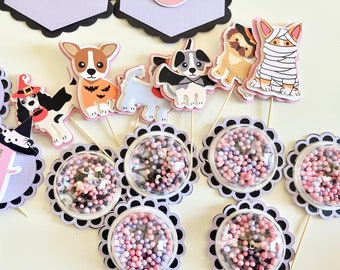 May include: Halloween-themed dog cupcake toppers and decorative elements. The toppers feature various dog breeds in costumes, including a witch, a bat, and a mummy. The image also includes decorative elements with a floral design and small, colorful beads.