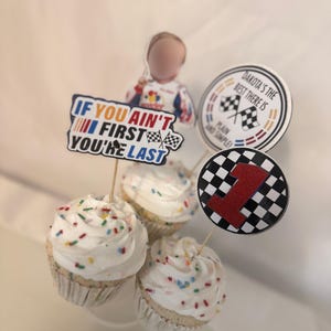 May include: Three cupcakes with white frosting and colorful sprinkles. Each cupcake has a decorative topper, including one with the text "If you ain't first you're last", a checkered flag, and a red number 1. The theme is racing.