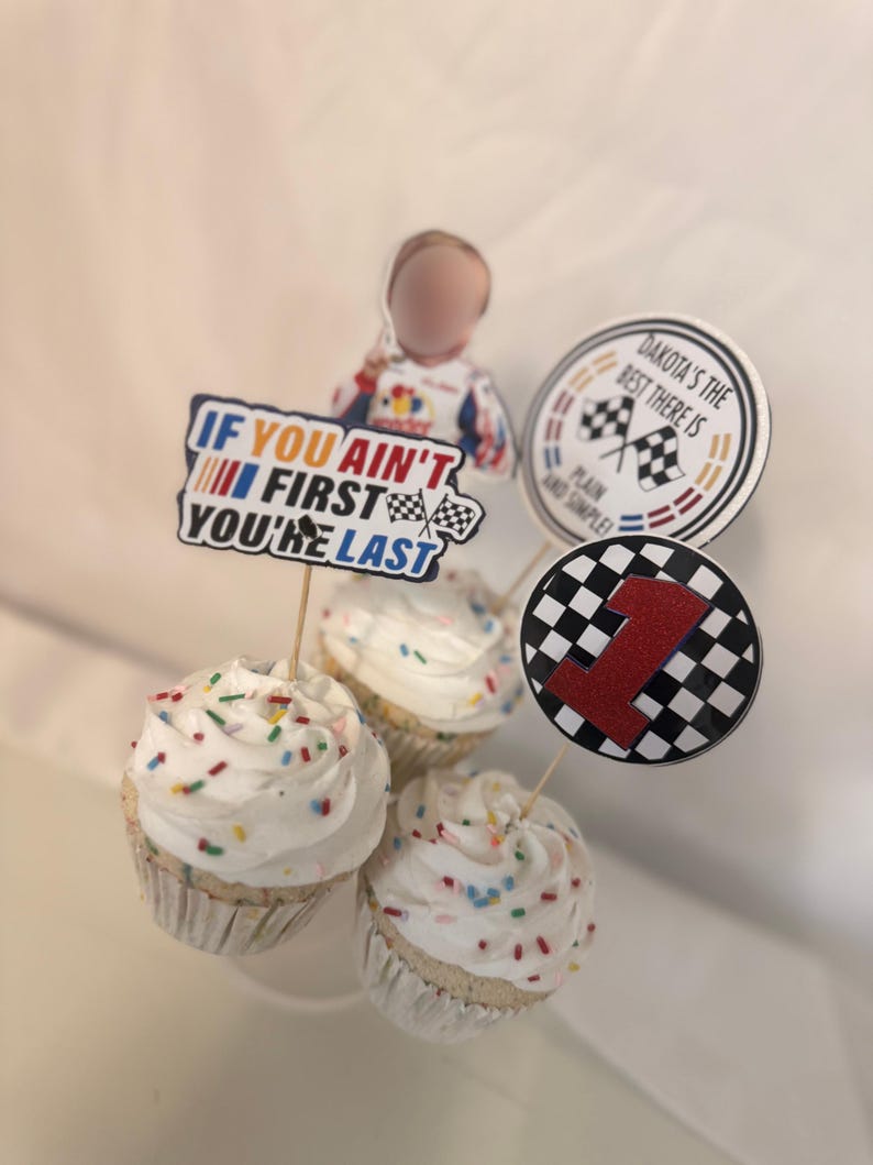 May include: Three cupcakes with white frosting and colorful sprinkles. Each cupcake has a decorative topper, including one with the text "If you ain't first you're last", a checkered flag, and a red number 1. The theme is racing.