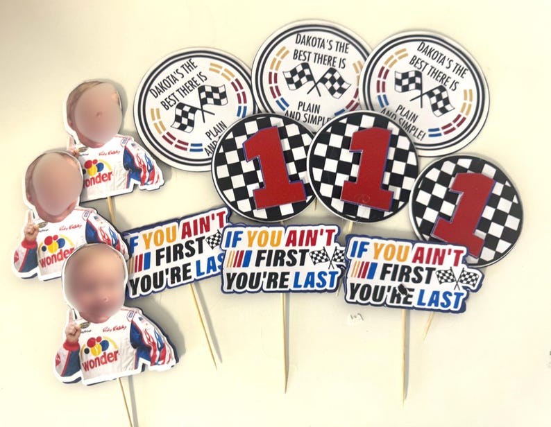 May include: A collection of racing-themed party decorations. Includes circular toppers with checkered flags and the text "Dakota's the best there is" and "1". Also, cutouts of race car drivers and signs that read "If you ain't first you're last".