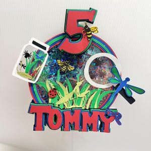 Può includere: Un cake topper a strati per compleanno con il numero 5 rosso, un barattolo con insetti e il nome "TOMMY" in lettere rosse. Il topper presenta una libellula, una coccinella e un'ape. Lo sfondo è un cerchio con strati blu, verdi e rossi.